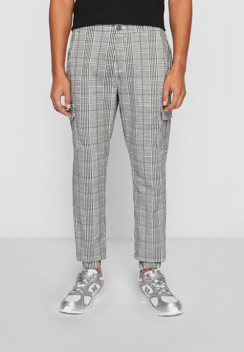 Homme portant un pantalon cargo à carreaux noir et blanc et des baskets New Balance argentées, debout devant un fond uni.