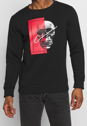 Sudadera negra con cuello redondo, que presenta un gráfico de calavera plateada dentro de un rectángulo rojo y texto estilizado en blanco. Tejido suave tipo jersey.