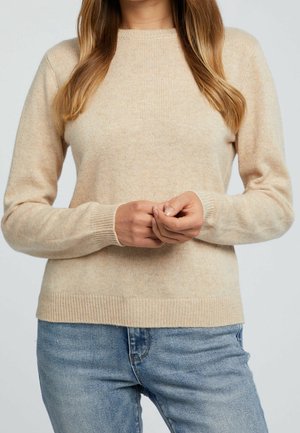 Femme aux longs cheveux châtain clair portant un pull beige et un jean bleu, debout les mains doucement jointes devant elle.