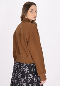 Jongeman met lang haar, gekleed in een bruin geribbeld jasje en een zwarte bloemenrok, kijkt over haar schouder tegen een effen achtergrond.