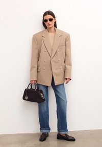 Naine kannab ülekaalu beezist triibulist bleiserit, siniseid teksaseid, musti leopardimustriga mokassine, prille ja hoiab musta käekotti.