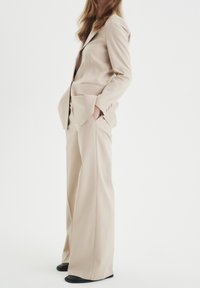 Valkoinen räätälöity puku, jossa on istuva bleiseri ja leveälahkeiset housut. Silkkinen kankaan pinta, yksirivinen malli ja etutaskut. Mustat jalkineet.