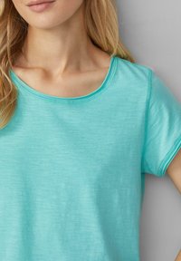 T-shirt à manches courtes de couleur teal clair avec un col rond et une texture douce et lisse. Des détails de couture le long des bords sont visibles.