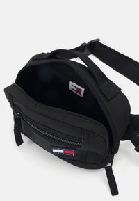 Melna auduma crossbody soma ar rāvējslēdzēja nodalījumu, regulējamu siksnu un redzamu logotipa etiķeti. Iekša ir bez raksta un gluda.