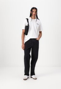 White cotton t-shirt with black text, relaxed-fit black denim jeans, and white sneakers with brown accents. Black crossbody bag on shoulder.