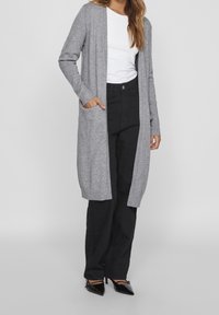 Long gilet gris en maille, avec des poignets et des poches côtelés, assorti d'une chemise blanche et d'un pantalon noir.