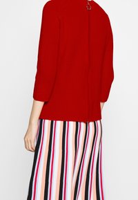 Roter Strickpullover mit rundem Ausschnitt und Reißverschluss auf der Rückseite, kombiniert mit einem mehrfarbigen gestreiften Rock. Texturiert.