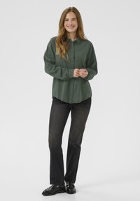 Dark green button-up shirt with long sleeves, worn over black jeans and black shoes. Features a relaxed fit and smooth texture.