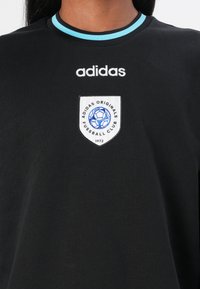 Sort bomulds t-shirt med rund halsudskæring og turkist kantbånd, "adidas" logo og et broderet klubmærke i skjoldform.