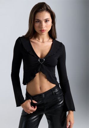 Sort langærmet cropped top med en flæset V-halsudskæring og hjerteformet sølvaccent, parret med sorte bukser i faux læder.