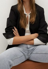 Blazer negro con mangas arremangadas, camiseta blanca con cremallera y jeans de mezclilla azul claro. Modelo sentado, con los brazos cruzados, cabello largo y ondulado. Collares sencillo con colgante.