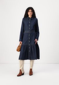 Robe en denim foncé avec un devant boutonné, un col et une jupe à volants. Assortie avec des bottines brunes et des collants à motifs. Petite sacoche marron.