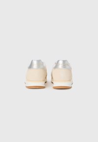 Une paire de baskets en suède beige avec des onglets de talon argentés et un logo doré en relief "Idée Sport Club" sur un fond blanc.