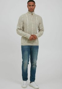 Beige kabelgebreide trui met hoge kraag en kwart-rits, gecombineerd met lichtblauwe jeans en witte sneakers.