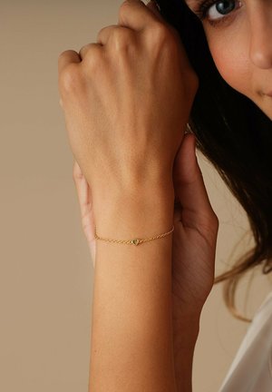 Poignet avec un délicat bracelet en chaîne d'or orné d'un petit pendentif en forme de cœur, tenu doucement par la main opposée sur un fond neutre.