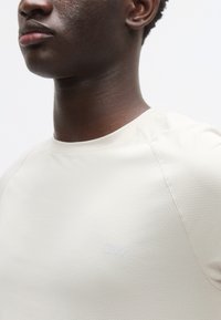 Μπεζ μπλουζάκι με υφή, στρογγυλή λαιμόκοψη και κοντά μανίκια, με ένα διακριτικό ανάγλυφο λογότυπο στο στήθος. Ομαλή υφή υφάσματος.