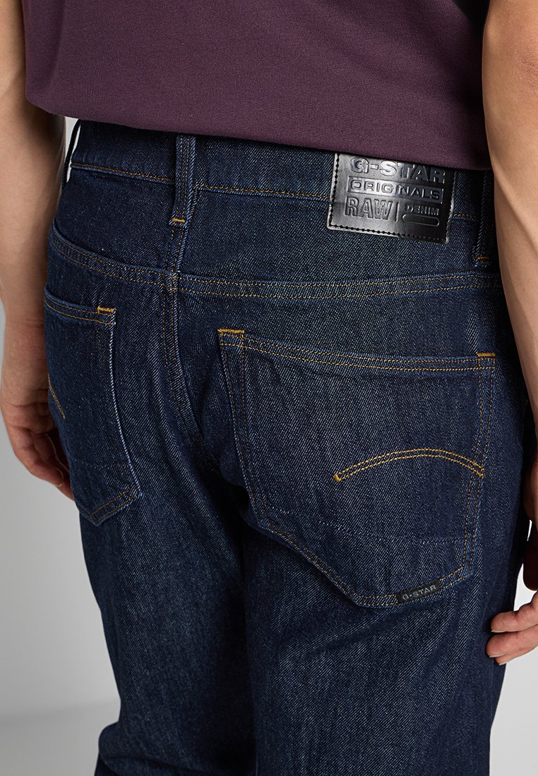 Jeans in denim blu scuro con cuciture gialle, cinque tasche e una etichetta in pelle nera sul cinturino posteriore.