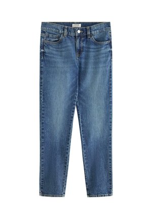 Dreng står med den ene hånd i lommen iført en hvid t-shirt, blå jeans og hvide sneakers mod en ensfarvet lys baggrund.