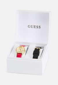 Goldfarbene Uhr mit rundem Ziffernblatt, rotem Lederarmband und einem mit Kristallen besetzten Gehäuse, begleitet von einem schwarzen Silikonarmband, in einer weißen Box.