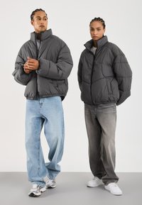 Graue Pufferjacken mit hohem Kragen, präsentiert an zwei Personen, die hell- und dunkelblau gewaschene, weite Jeans tragen, kombiniert mit Sneakers.