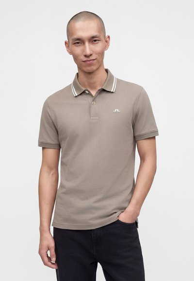 Homme à la tête rasée portant un polo taupe avec un col rayé blanc et un pantalon noir, debout avec une main dans la poche.