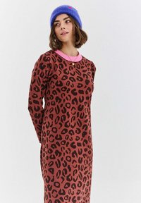 Žena oblečená v hnědé leopardí šaty s růžovým límcem a modrou pletenou čepicí, stojící před jednoduchým světlým pozadím.