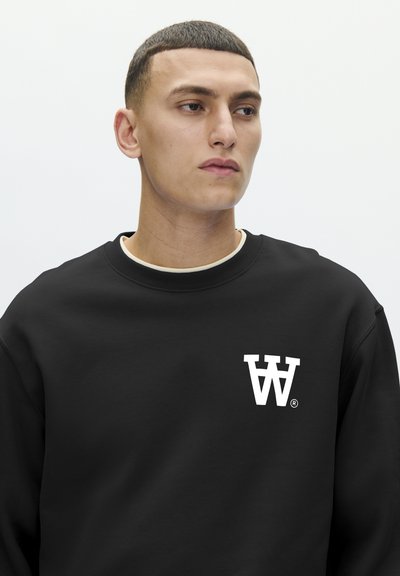 Sweatshirt noir avec un col rond, arborant un logo blanc sur le côté gauche de la poitrine. Le tissu semble doux et confortable.