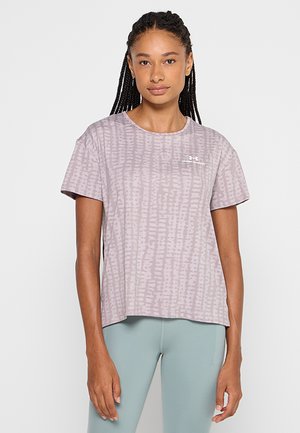 Femme aux longs cheveux tressés portant un T-shirt ample violet clair à motifs et des leggings bleu clair, debout devant un fond uni.