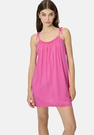 Joven mujer con un vestido mini sin mangas de color fucsia brillante con tirantes de lazo, posando frente a un fondo liso.