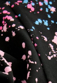 Black fabric with irregular pink and blue paint splatter patterns, shown in close-up folds and texture details.