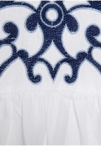 Weißer Stoff mit blauen gestickten Blumen- und Wirbelmustern. Ausgestattet mit detaillierter Stickerei und einem fransigen unteren Saum.
