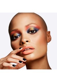 Bald individual with bold eye makeup featuring orange, blue, and gold shades, nude lips, and dark polished nails. Smooth skin texture.