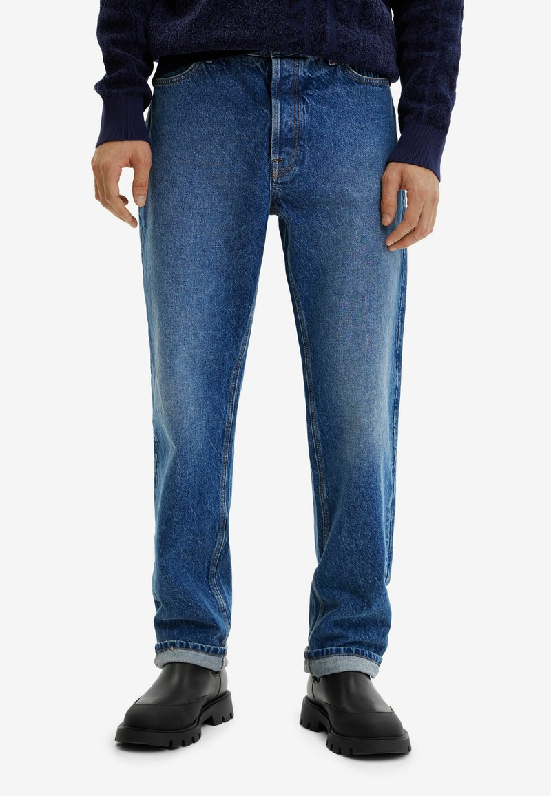 Homme portant un jean droit bleu avec des ourlets roulés, des bottes noires imposantes et un pull bleu foncé, se tenant devant un fond blanc.