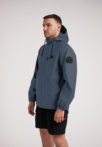 Chaqueta impermeable de color azul oscuro con capucha, puños elásticos, bolsillos frontales y un parche negro con el logo en la manga izquierda.
