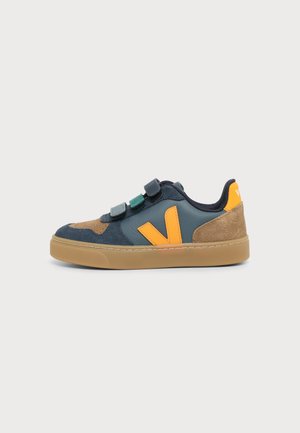 Zapatilla baja con ante marrón, piel azul oscuro, logo naranja y tres tiras de velcro azul-verde en suela de goma natural.