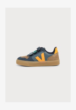 Zapatilla baja con ante marrón, piel azul oscuro, logo naranja y tres tiras de velcro azul-verde en suela de goma natural.