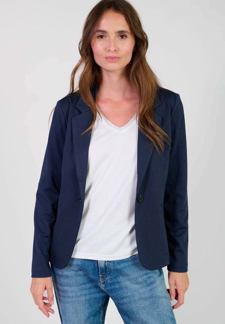 Blazer blu navy con un bottone, collo a sciarpa e tessuto morbido. Abbinato a una maglietta bianca a scollo a V e jeans azzurri chiari.