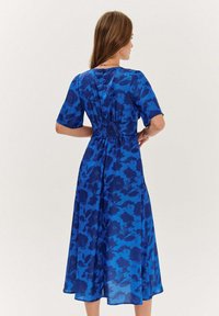 Modré šaty s květinovým vzorem, krátkými rukávy, nařaseným pasem a splývavou sukní. Tkanina má jemnou, lehkou strukturu.