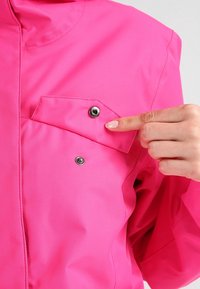 Ruka osoby ukazuje na otevřenou klopu kapsy se snap-button na jasně růžové bundě.