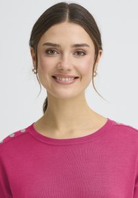 Pull en maille fuchsia avec un col rond, doté de boutons argentés sur les épaules et d'une texture douce et lisse.