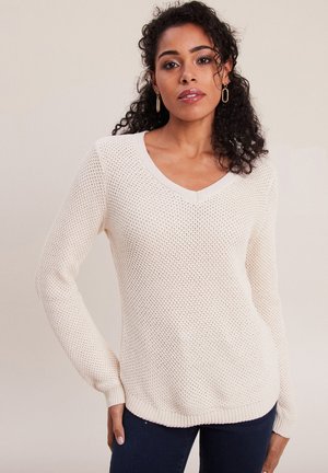 Light beige knit sweater with a v-neck, long sleeves, and a loose, textured weave. Hem is ribbed, providing a relaxed fit.