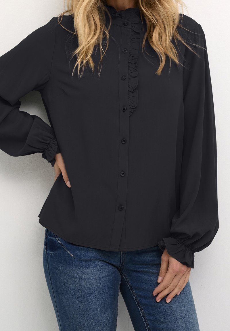 Blouse noire à manches longues avec un devant volanté, fermeture à bouton et poignets froncés. Texture lisse et coupe décontractée, associée à un jean bleu.