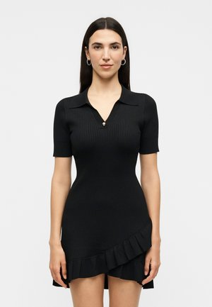 Γυναίκα με μακριά σκούρα μαλλιά που φοράει εφαρμοστό μαύρο ριμπ φόρεμα με γιακά, κοντά μανίκια, λεπτομέρειες με κουμπιά και βολάν σε ασύμμετρο τελείωμα.