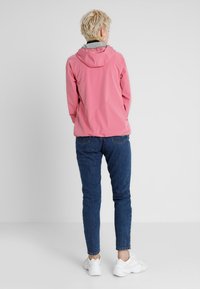 Lichtroze waterdichte jas met een capuchon, voorzien van een grijze voering, gecombineerd met blauwe jeans en witte sneakers.