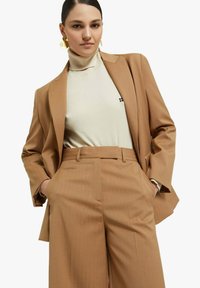 Blazer marrone a righe sottili abbinato a un dolcevita crema, completato da pantaloni coordinati. Presenta una vestibilità comoda e una texture liscia. Orecchini dorati visibili.