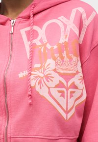 Κοντινό πλάνο από ένα ροζ φούτερ με φερμουάρ, με την επιγραφή «ROXY», λουλουδένια σχέδια, μια κορώνα και πλεκτά κορδόνια.