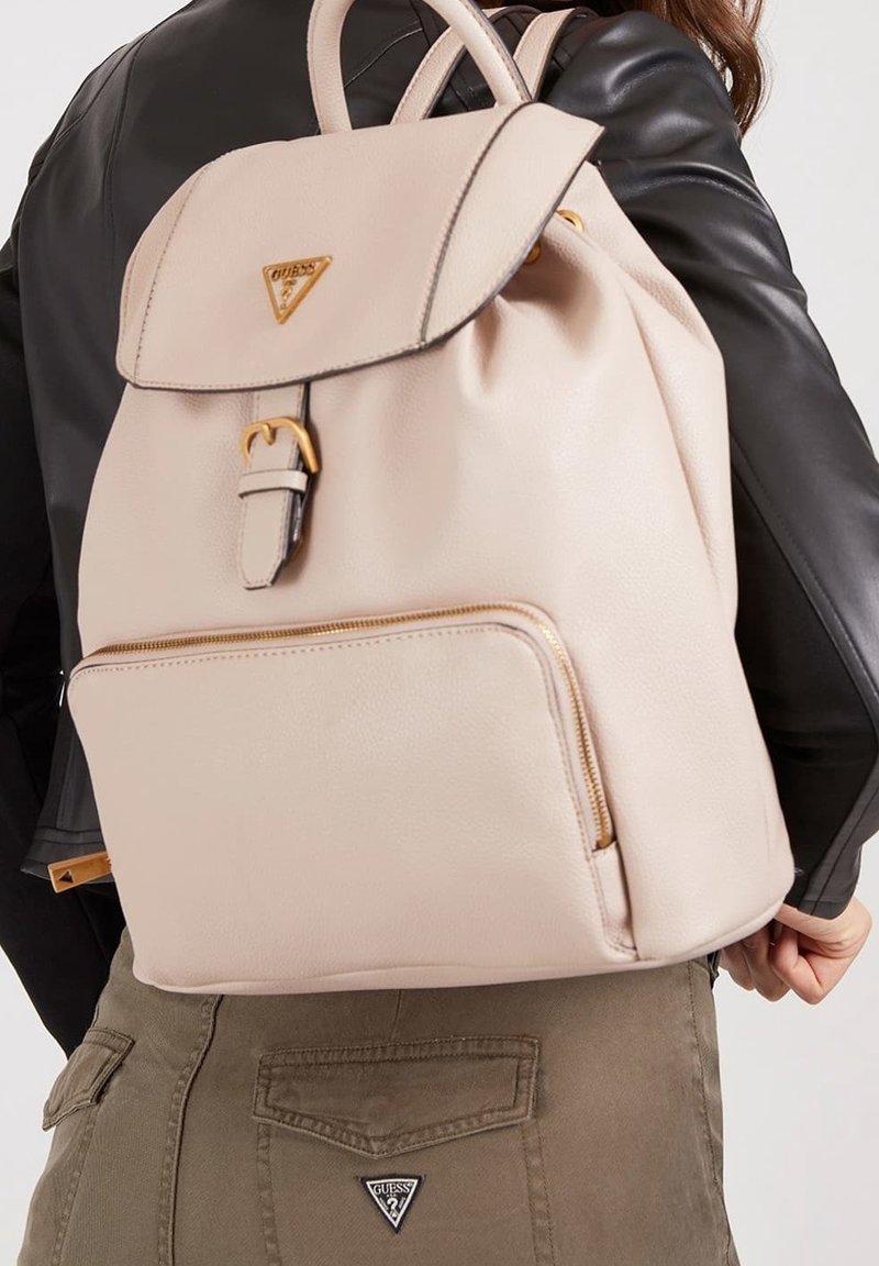 Personne portant un sac à dos Guess beige avec une poche avant zippée, une boucle dorée et un logo sur le rabat, vêtue d'une veste noire et d'un pantalon marron.