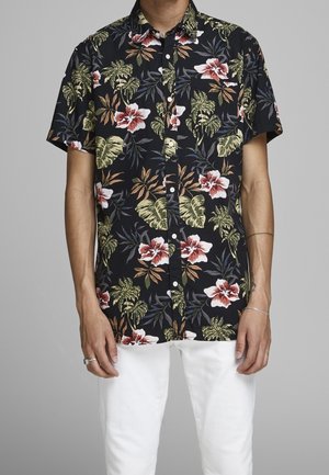 Homme portant une chemise noire à manches courtes avec boutons, ornée de fleurs d'hibiscus rouges et blanches ainsi que de feuilles tropicales vertes, associée à un pantalon blanc.
