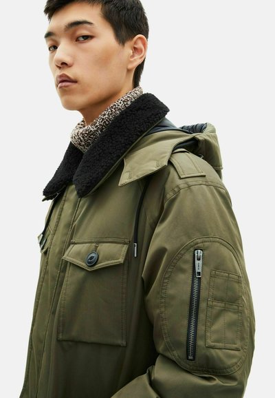 Zielona oliwkowa parka z czarnym kołnierzem wykończonym futrem, licznymi kieszeniami i zamkami błyskawicznymi. Posiada trwały materiał i militarny styl.