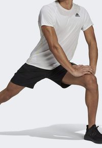 Witte sportieve shirt met korte mouwen van textuurstof, gele accenten, gecombineerd met zwarte shorts. Model staat in een lungespositie en draagt zwarte sneakers.